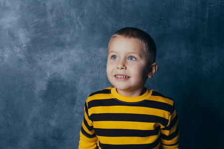 Little fashionable boy posing in front of a gray-blue concrete wall. Portrait of a smiling child dressed in a black and yellow striped sweater and brown pants.の写真素材