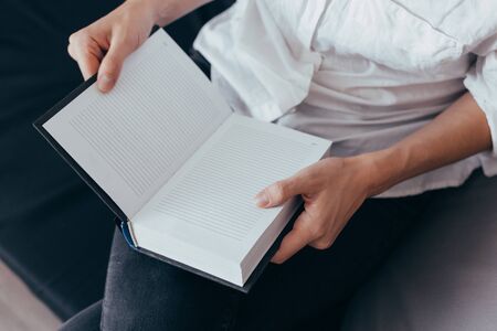 Beautiful female hands hold an open book or magazine in a room on a black sofaの写真素材