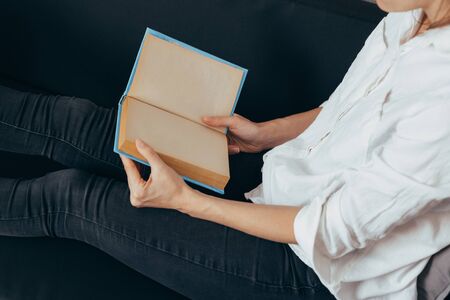 Beautiful female hands hold an open book or magazine in a room on a black sofaの写真素材