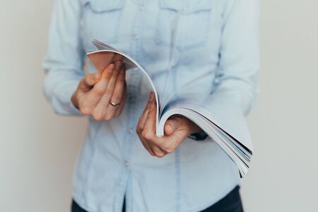 Beautiful female hands are holding an open book or magazine in a room on against a light wallの写真素材