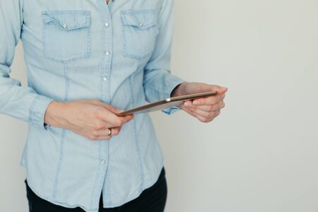 Image of a young woman in a blue shirt working on a tablet computer at home on a background of light wallの写真素材