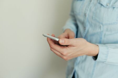 Image of a young woman in a blue shirt working on a modern touchscreen phone at home against a light wallの写真素材