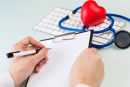 doctor's hands in a white medical coat write a prescription, white keyboard and red heart on a blue background. concept of  cardiologyの写真素材