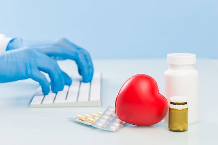 Doctor's hands in blue medical gloves are typing on a white keyboard, next are pills and a red heart...