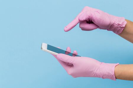 Female hand in a pink medical glove holds a blank modern mobile phone on a blue background. Mock upの写真素材