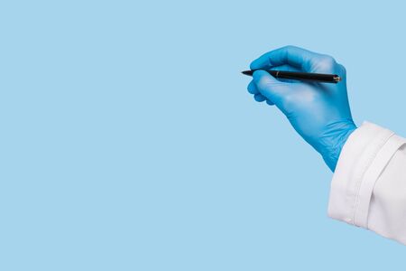A female hand in a medical glove and in a white coat holds a pen and writes in the air on a blue background.の写真素材