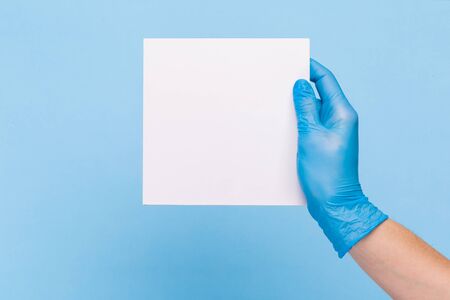 female hand in a sterile medical glove holds a blank form of paper on a blue background. Mock up, business card, flyerの写真素材