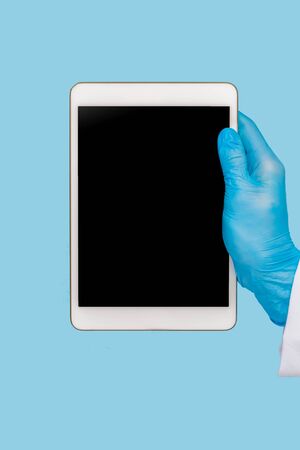 Female hand in a  medical glove holds a blank modern tablet on a blue background. Mock up.の写真素材