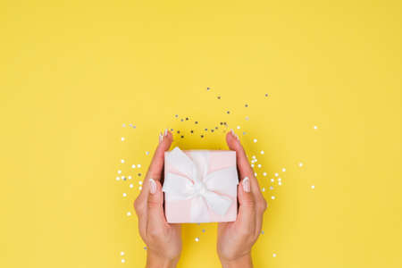 Women hands holding a gift or gift box decorated with confetti on a yellow background top view. Flat composition for birthday or wedding.の写真素材