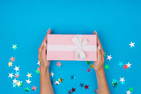 Women hands holding a gift or gift box decorated with confetti on a blue table top view. Flat composition for birthday or wedding.の写真素材