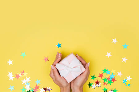 Women hands holding a gift or gift box decorated with confetti on a yellow background top view. Flat composition for birthday or wedding.の写真素材