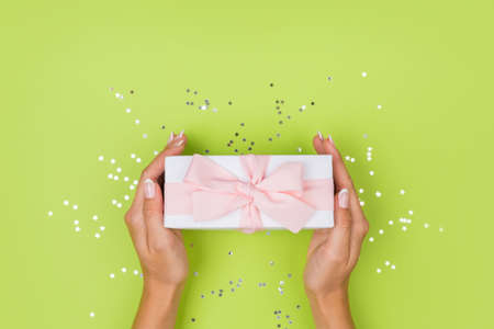 Women hands holding a gift or gift box decorated with confetti on a green table top view.