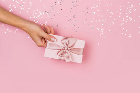 Women hands holding a gift or gift box decorated with confetti on a pink pastel table top view. Flat composition for birthday or wedding.の写真素材