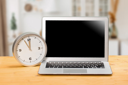 Laptop with blank screen, clock with arrows at five to twelve on a wooden table. mockup for your textの写真素材