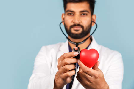 Cute modern Indian / Asian doctor with a stethoscope and a red heart in his hands, in a white coat...