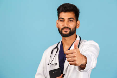 Handsome modern indian / asian doctor with stethoscope, in uniform writing prescription on tablet on...