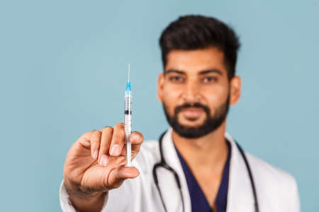 Cute modern Indian / Asian doctor with a stethoscope and a syringe in his hands, in a white coat on a blue background. space for textの写真素材