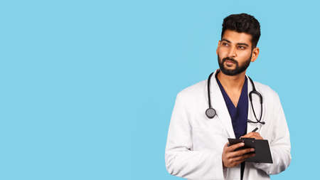 Handsome modern indian / asian doctor with stethoscope, in uniform writing prescription on tablet on blue background medicine, healthcareの写真素材