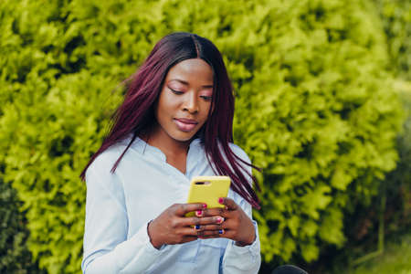 smiling african american girl with pink hair and black hat sitting on a bench with a mobile phone...