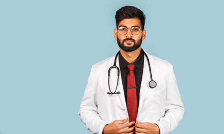 Cute modern Indian / Asian doctor wearing glasses with stethoscope, tie and white coat on light blue background. space for textの写真素材