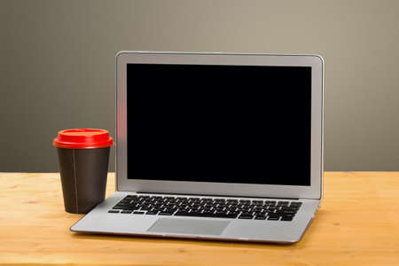 A laptop with a blank screen for product display and a cup of coffee on a wooden table and a plain wall. mockupの写真素材
