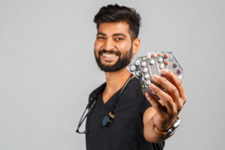 amusing young indian doctor or surgeon with stethoscope and pills on gray backgroundの写真素材
