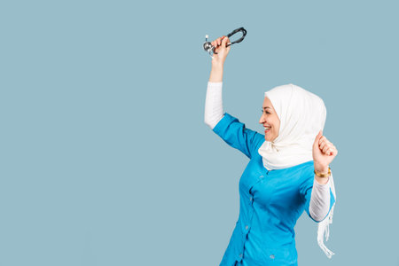 Portrait of a friendly, muslim doctor or nurse woman in hijab with a stethoscope on a blue background. emotions of victory or successの写真素材