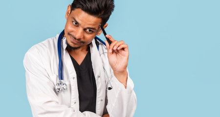 handsome young Indian / Asian doctor with a stethoscope, in a white coat, is poking about a problem on a blue background. new diseasesの写真素材