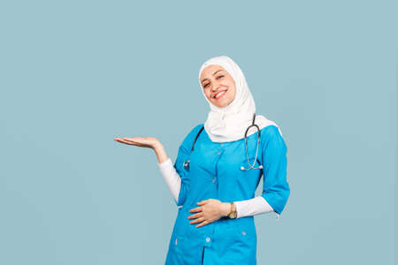 Portrait of friendly, muslim doctor or nurse woman in hijab with stethoscope pointing to blank space...