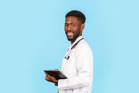 African american male doctor with stethoscope in white coat writing prescription on blue backgroundの写真素材
