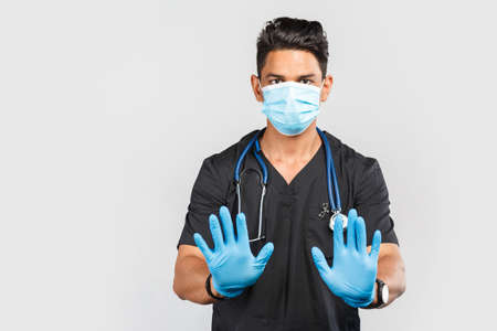 Handsome modern Indian / Asian doctor with stethoscope wearing blue medical face mask and gloves on gray background.の写真素材