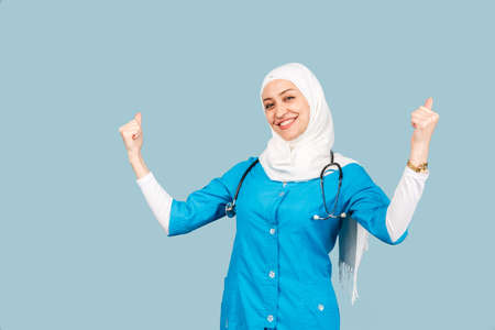 Portrait of a friendly, muslim doctor or nurse woman in hijab with a stethoscope on a blue background. emotions of victory or successの写真素材