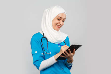 Portrait of a friendly, Muslim doctor or nurse woman in a hijab with a stethoscope writes a prescription on pills on a gray background.の写真素材