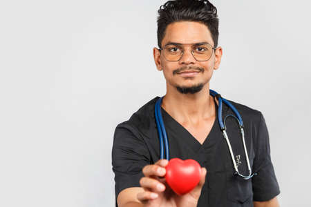 young Indian / Asian doctor with stethoscope in uniform on a gray background holding a red heart in...