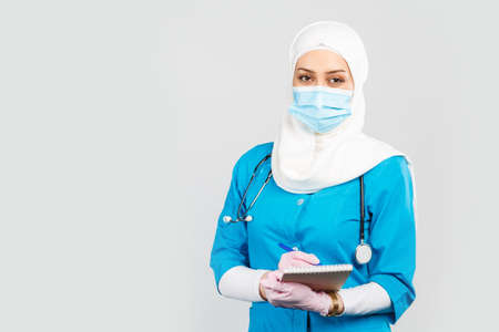Portrait of friendly muslim doctor or nurse wearing hijab and medical face mask with stethoscope writing prescription to patient on gray background.の写真素材