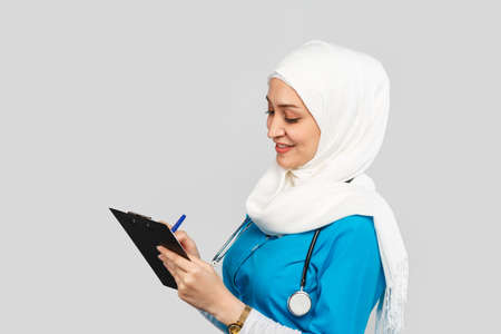 Portrait of a friendly, Muslim doctor or nurse woman in a hijab with a stethoscope writes a prescription on pills on a gray background.の写真素材