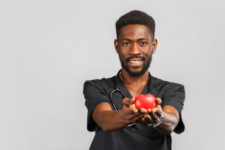 cute african doctor cardiologist with stethoscope holding red heart in hand while standing against...