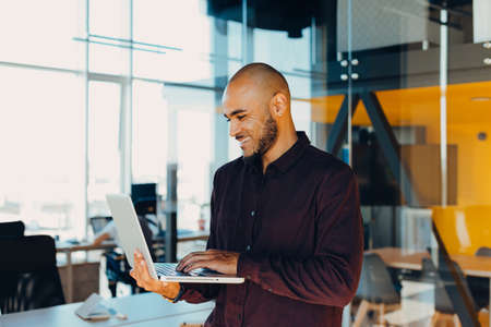 Smiling bearded african man using laptop at home, sitting on a wooden table. Concept of young people...