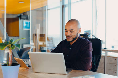Smiling bearded african man using laptop at home, sitting on a wooden table. Concept of young people...