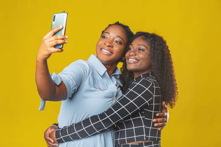 two friends beautiful african american girls take a selfie or video call with friends on a yellow backgroundの写真素材