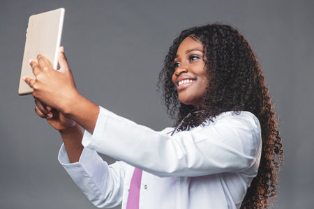 happy professional doctor black woman in white lab coat with stethoscope and tablet pc online consultations for her patientsの写真素材