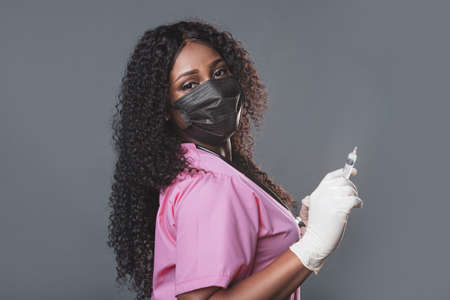 close-up portrait of a young african american woman doctor in a mask with gloves and a syringe in her hands on a gray background. medical worker posing on light background. Pandemia conceptの写真素材