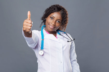 African woman doctor in a white coat and with a stethoscope shows a sign of approval with her hands, thumbs up, okay.の写真素材