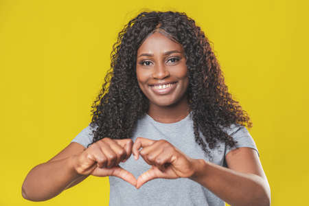 Beautiful african american girl with afro hairstyle smiling and showing with hands a heart sign with...