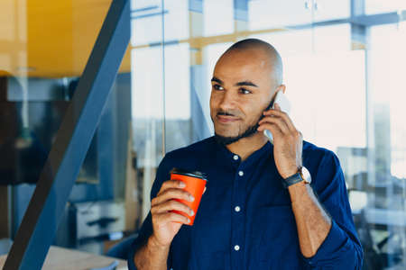 smiling young black designer creative man posing in workspace with a glass of coffee in his handsの写真素材