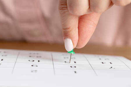 a woman's hand holds a green button to mark the date on a white calendar on the tableの写真素材