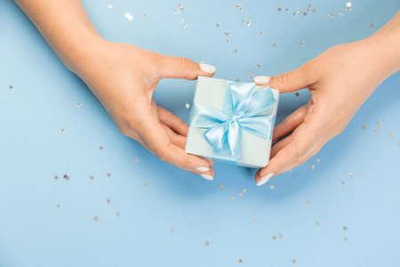 Flat lay of women's hands holding a gift wrapped and decorated with a bow on a blue background with...