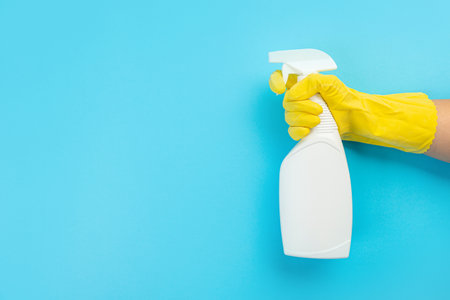A hand in a yellow glove holds a white bottle of detergent on a blue background. Banner with copy space. Chemical cleaners, household chemicals, brushes and collage supplies.の写真素材