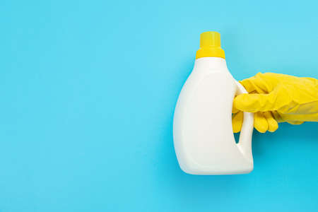 A hand in a yellow glove holds a white bottle of detergent on a blue background. Banner with copy space. Chemical cleaners, household chemicals, brushes and collage supplies.の写真素材