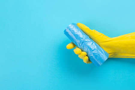 A hand in a yellow glove holds a plastic disposable trash bag on a blue background. place with copy...